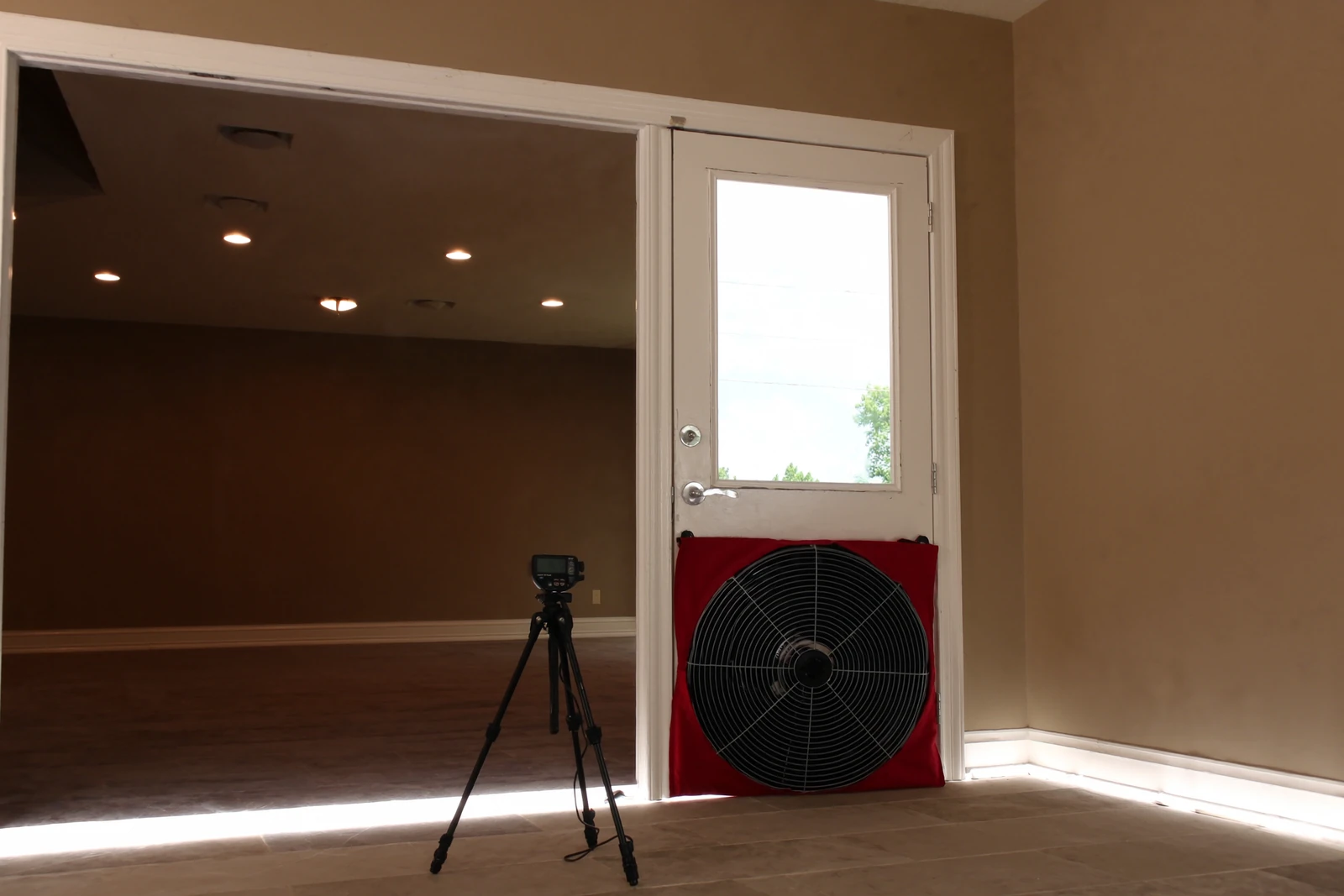 Blower door fan in a red panel beside a doorway with a gauge on a tripod